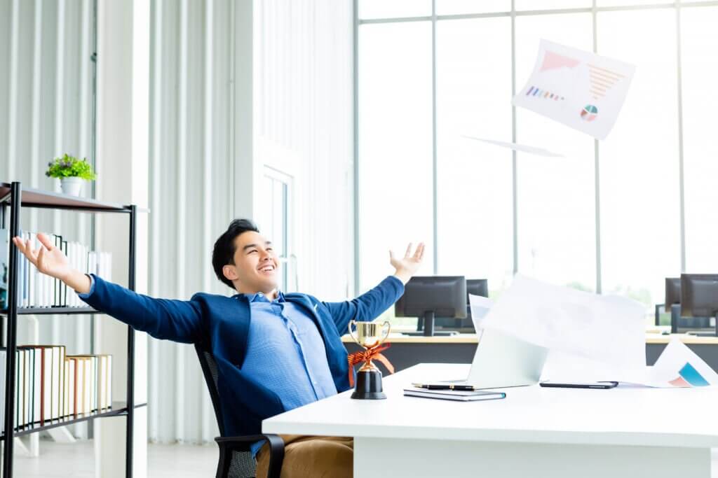 A young man in a suit celebrates with arms outstretched in an office, papers flying around him. A trophy sits on his desk, symbolizing success.