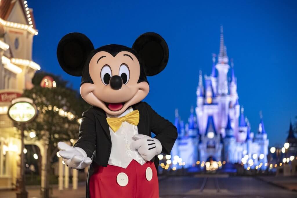 Mickey Mouse stands on Main Street, U.S.A. at Magic Kingdom® Park, with Cinderella Castle illuminated in blue at dusk — Walt Disney World® Resort.
