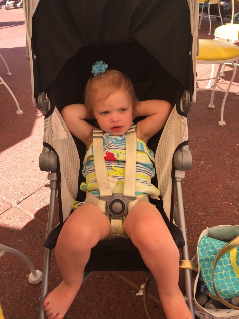 A toddler leaning back in a stroller in the shade at a theme park on a hot afternoon